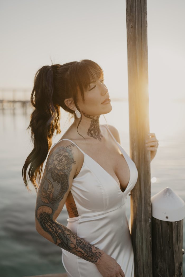 Bride Corrine posing on a dock at sunset during her waterfront wedding, wearing a white satin gown with the sun reflecting across the water.